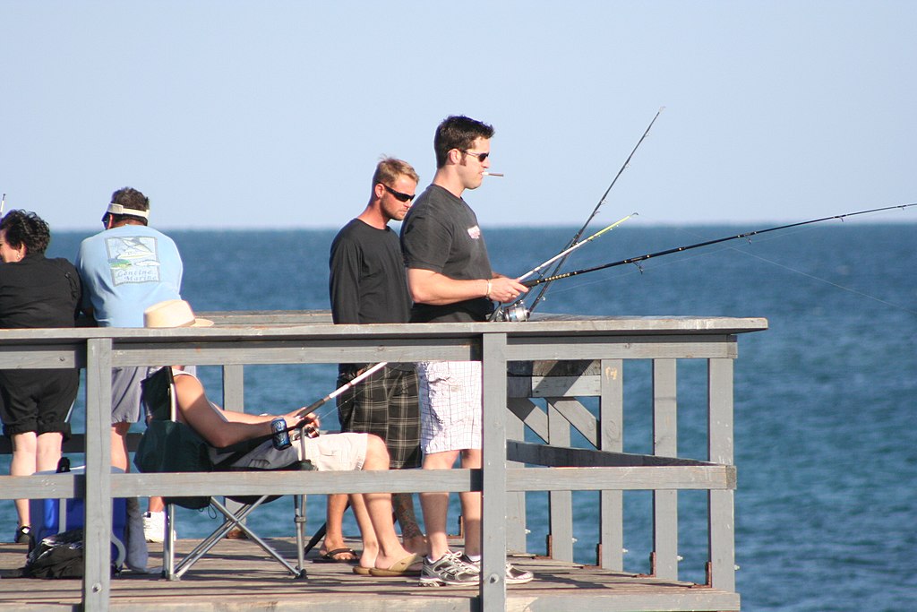 Four people fish from a wooden pier under a sunny, clear blue sky. One person relaxes on a chair, while others stand, holding fishing rods.