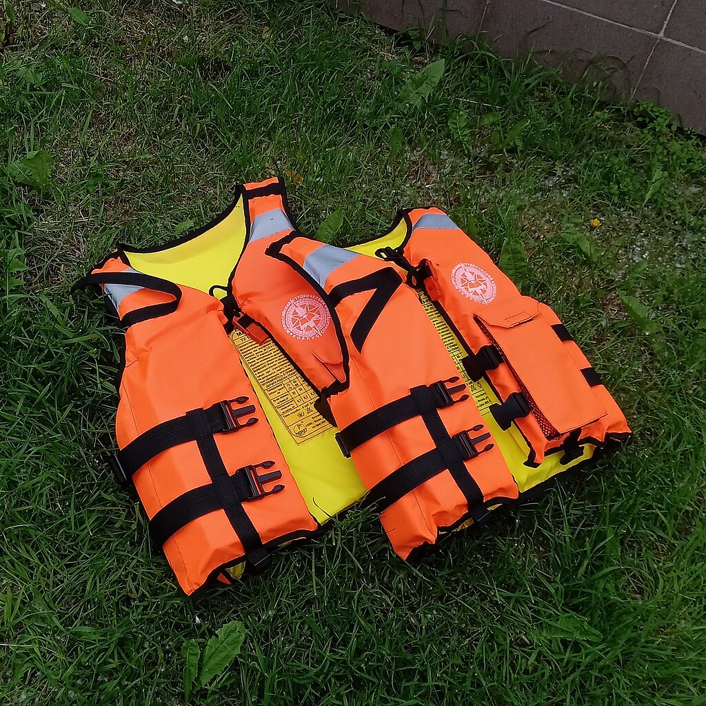 Two bright orange life jackets with black straps and reflective patches lie on green grass. The scene conveys readiness and safety awareness.
