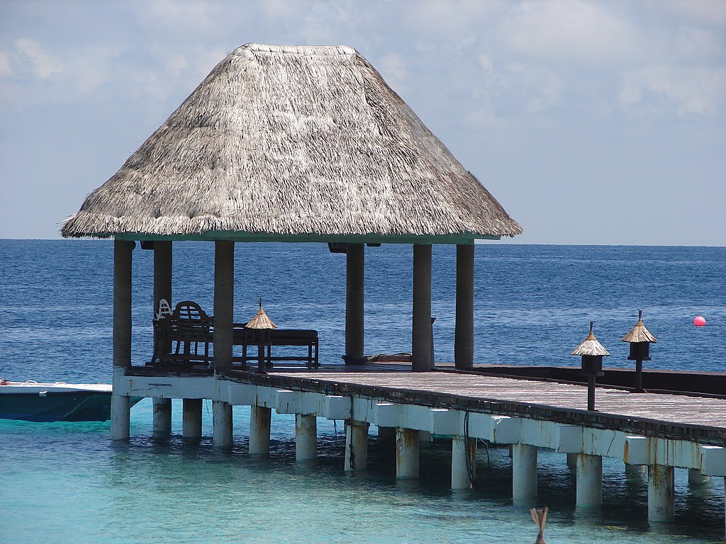 Thatch-roofed gazebo on a wooden pier over clear blue water, with a distant horizon. Calm and serene coastal setting under a lightly clouded sky.