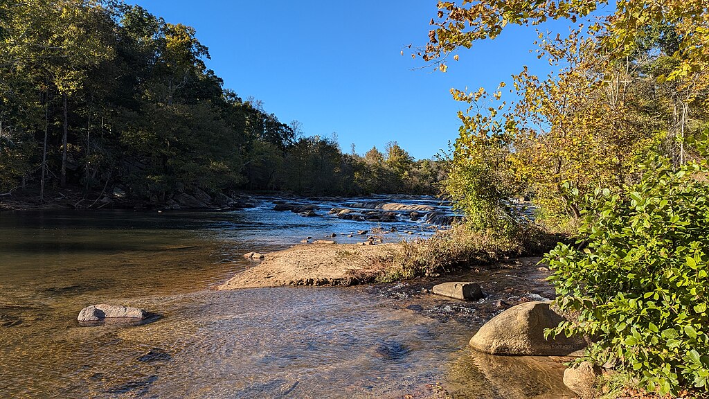 Calm river scene with clear blue sky, surrounded by lush green and autumn-colored trees. Rocky shoreline with gentle water flow creates a serene atmosphere.