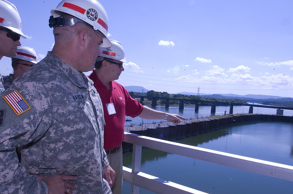 Three men in hard hats stand on a bridge overlooking a river and dam. One, in military uniform, listens as another points out features. Bright, clear day.
