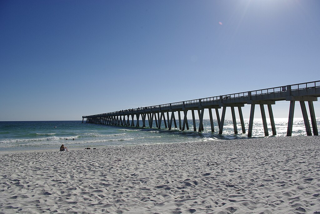 Sunny day at a beach with white sand and a long pier extending into the sparkling turquoise ocean. A person relaxes on the sand, conveying tranquility.