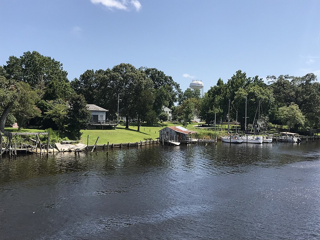 Peaceful riverside scene with boats docked near a small pier, surrounded by lush trees and quaint houses under a clear blue sky. Calm, serene atmosphere.