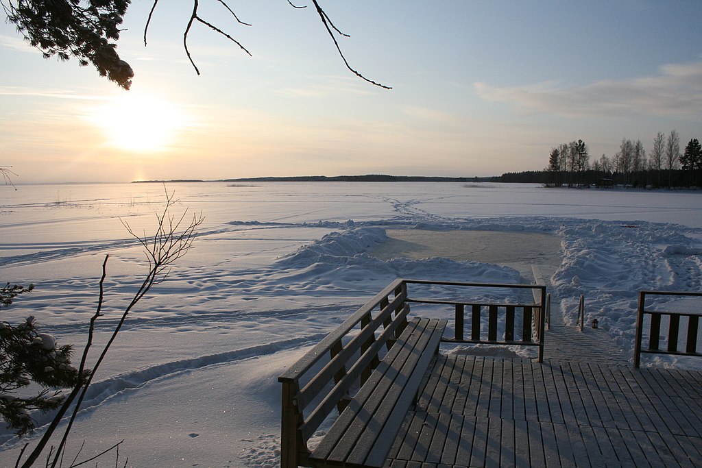 Sunset over a frozen lake with a wooden dock in the foreground. Snowy landscape, bare trees, and a serene, cold atmosphere.