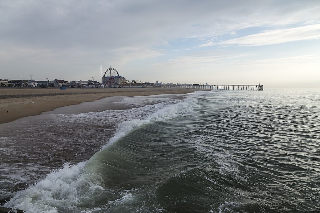 A serene beach scene with gentle waves rolling onto the shore under a cloudy sky. A distant Ferris wheel and a long pier stretch into the calm sea.