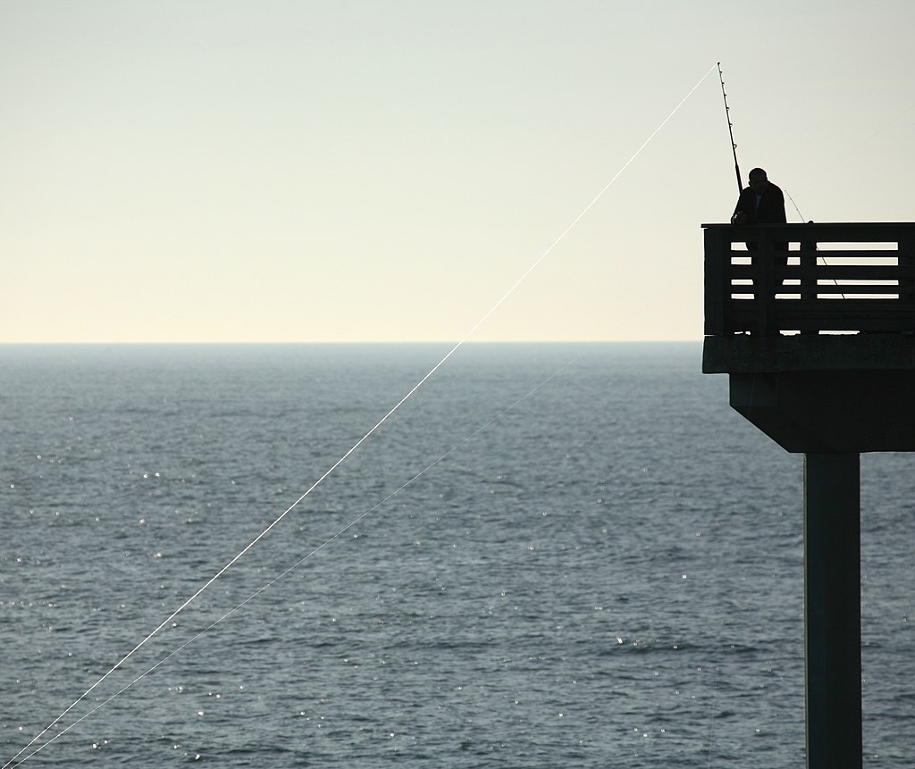 Silhouette of a person fishing off a pier against a vast, calm ocean under a clear sky. The scene conveys tranquility and solitude.