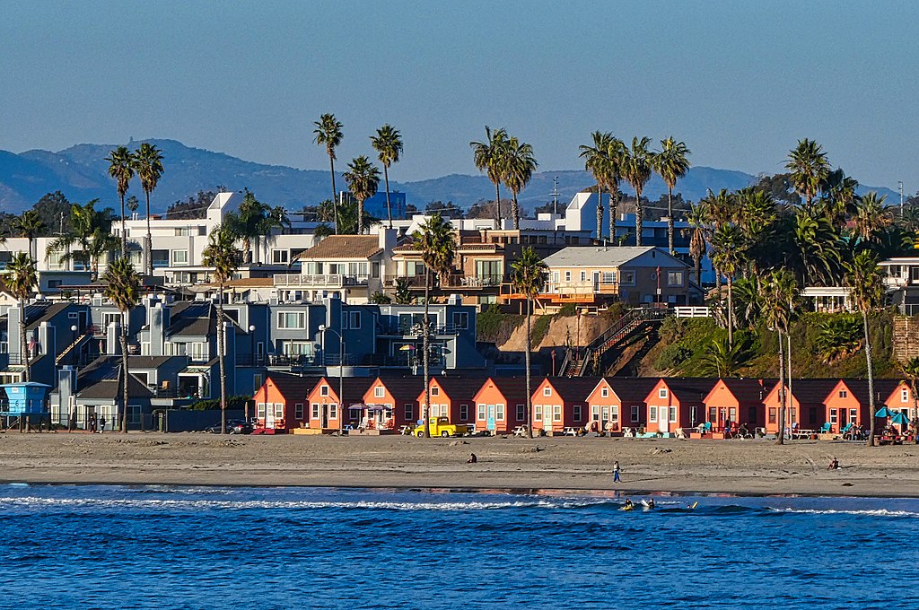 A row of red beach huts and modern houses behind palm trees line a sandy beach under a clear blue sky, with distant mountains in the background.