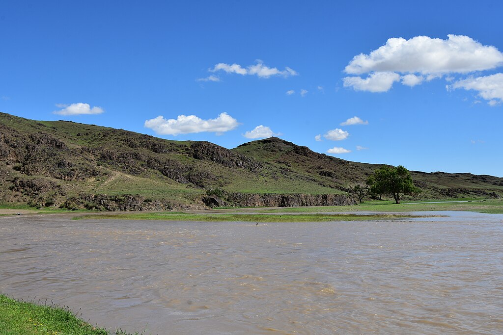 A wide river flows in the foreground with green hills and rocky terrain in the background under a clear blue sky with scattered fluffy clouds.