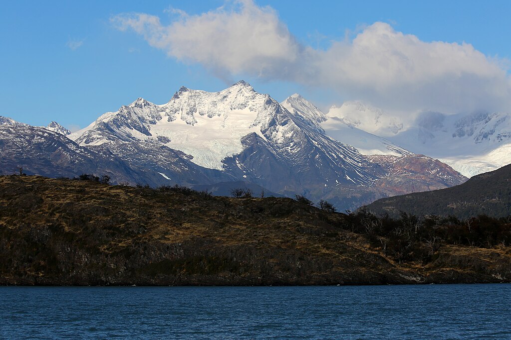 Snow-capped mountains under a blue sky with clouds, behind a rocky, forested hill. A calm, blue lake stretches across the foreground. The scene is serene and majestic.