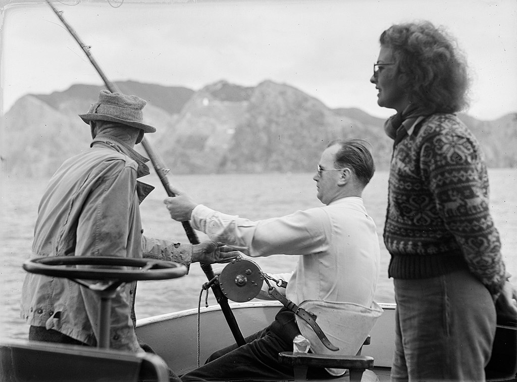 View of two men, and a woman on-board a fishing boat. The woman is wearing a patterned woollen jersey. One of the men is seated, and is holding a fishing rod. A body of water, and an island, is in the background.