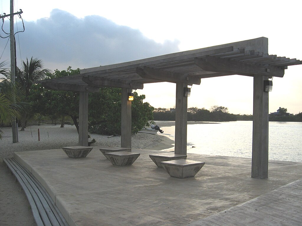 Concrete pergola with three benches on a beachside patio at sunset, with palm trees and shoreline in the background. The atmosphere is calm and serene.
