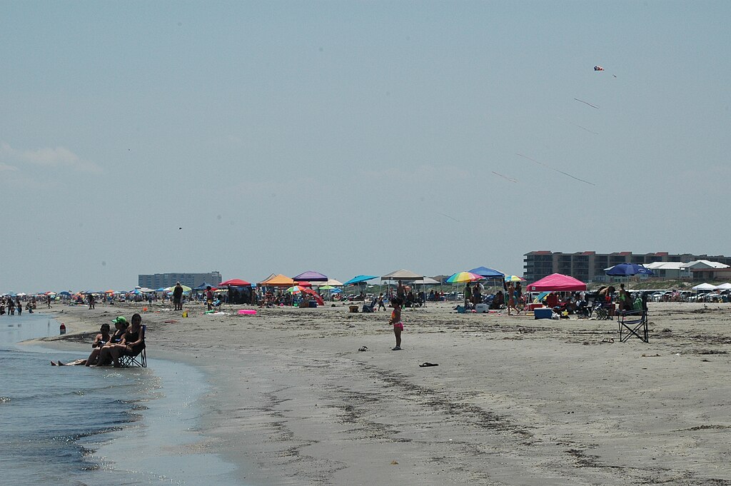 Crowded beach scene with colorful umbrellas and groups of people relaxing on the sand. Calm water to the left and buildings in the background under a clear sky.