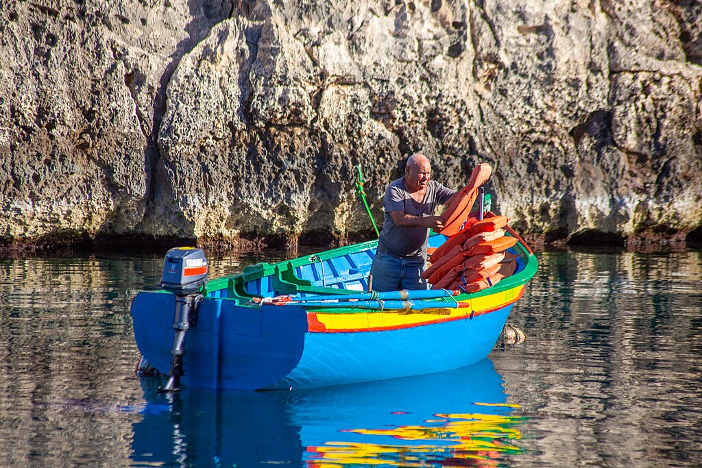 A man stands on a vibrant blue and yellow boat, organizing orange life jackets. The calm water reflects the rocky cliff behind, conveying a serene mood.
