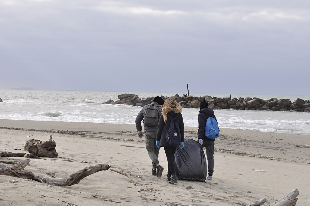Three people walk along a cloudy beach carrying a large trash bag, engaged in cleanup. Waves crash against rocks, creating a sense of community effort and care.