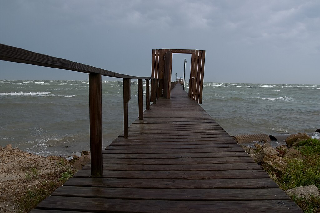 Wooden pier extending over choppy, gray sea under cloudy sky, conveying a moody and contemplative atmosphere. Smooth railing lines draw the eye onward.