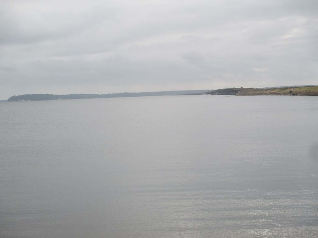 Calm gray sea under an overcast sky, with a distant shoreline and a bridge stretching across the water. The scene is tranquil and serene.