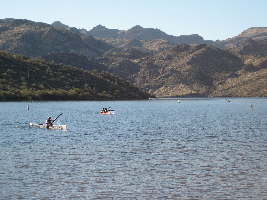 Kayakers paddle across a serene lake surrounded by rugged, sunlit mountains. The atmosphere is peaceful and adventurous, with a clear blue sky.