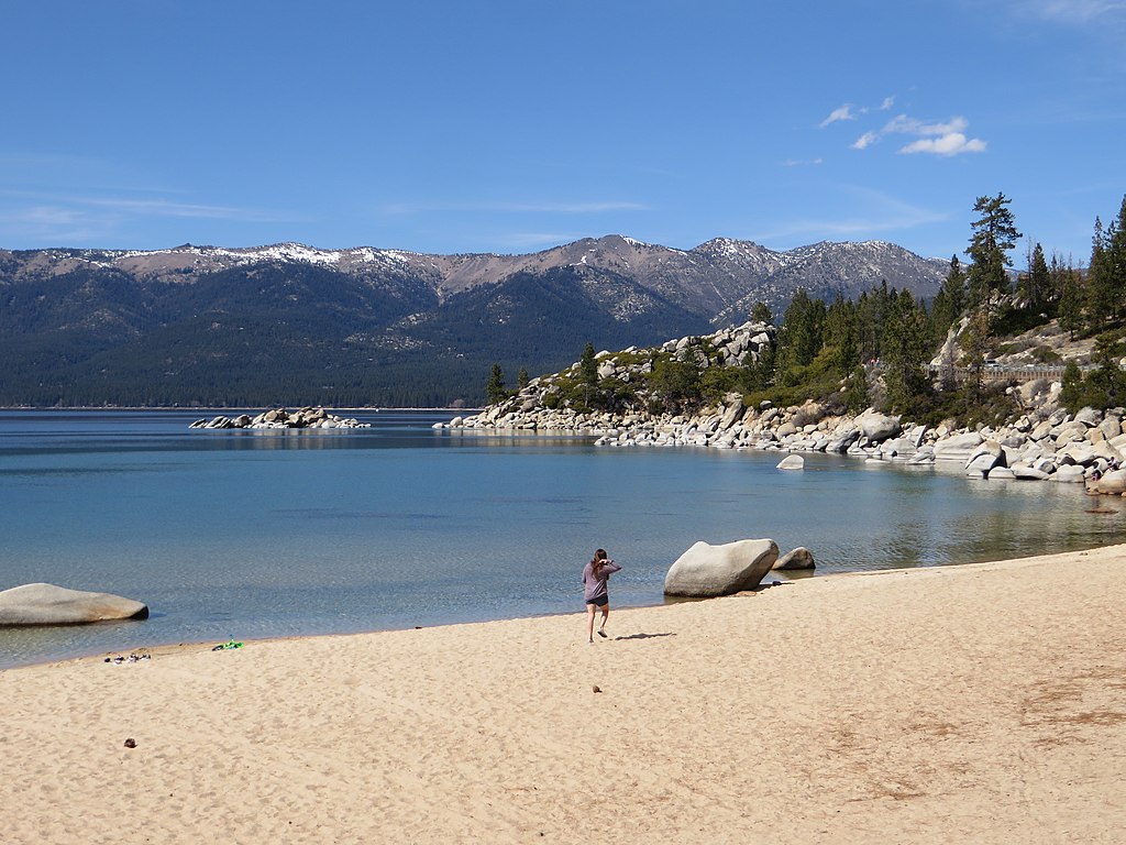 A person walks along a sandy beach beside a calm, clear lake, with rocky shoreline and snow-dusted mountains under a clear blue sky, evoking tranquility.