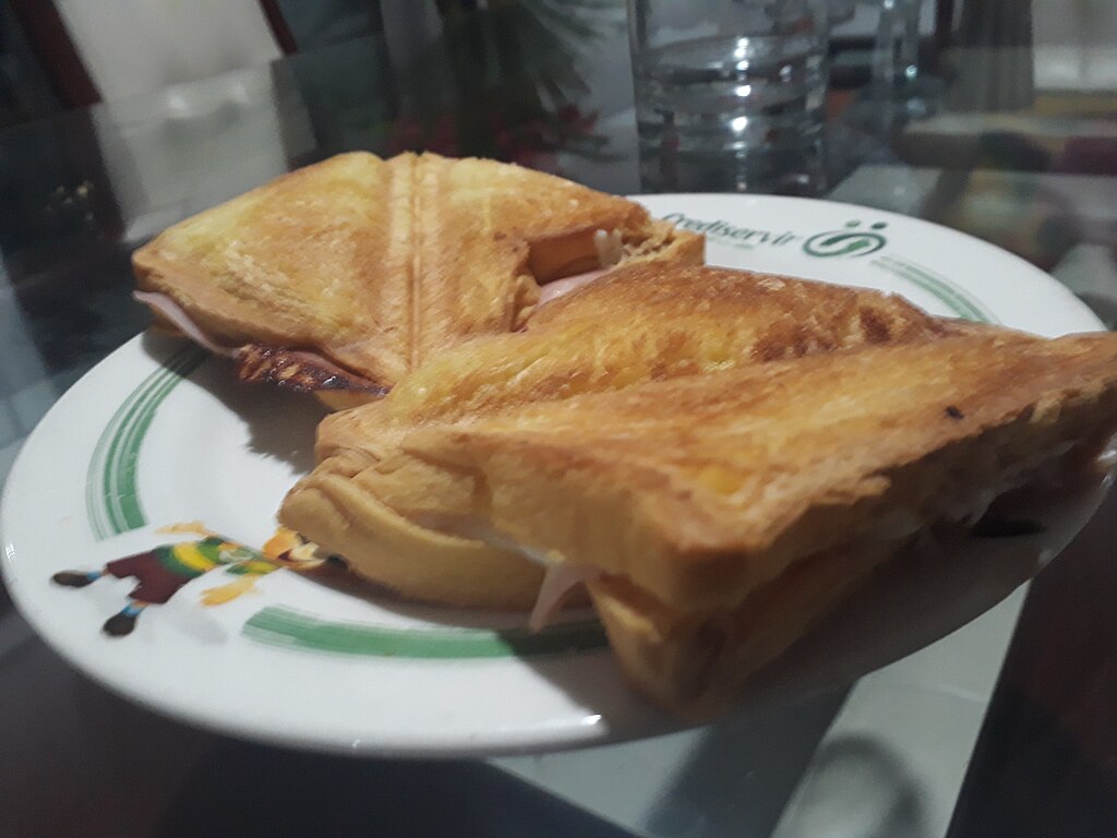Two golden-brown toasted sandwiches with visible ham and cheese on a white plate with a green stripe, placed on a glass table. Cozy and appetizing.
