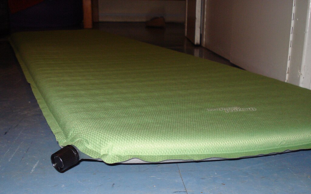 Green self-inflating camping mat on a blue floor in a dimly lit room. The mat is inflated, with a visible black valve near the edge.