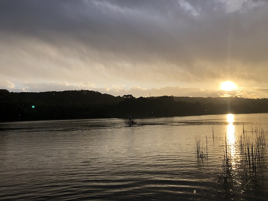 Sunset over a tranquil lake with a silhouetted tree line in the distance. The sky is a mix of golden and gray hues, reflecting on the water.