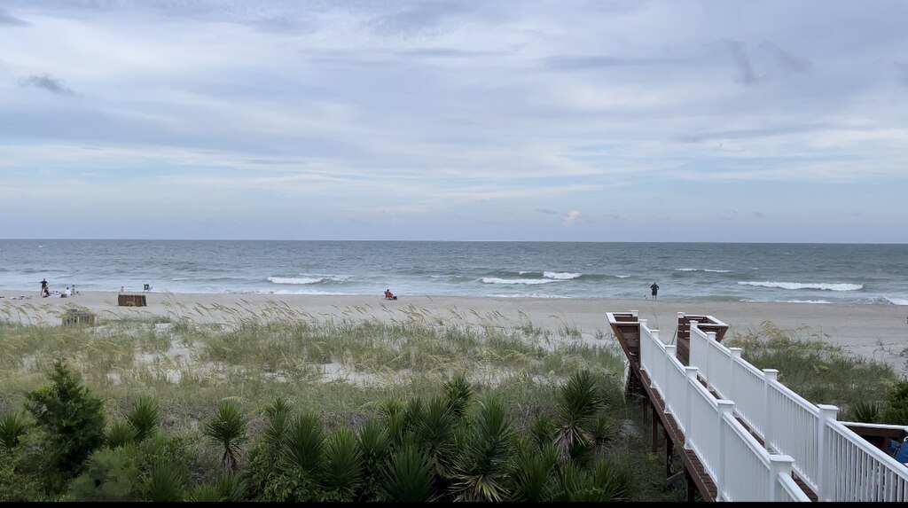 A serene beach scene with gentle waves under a cloudy sky. A wooden walkway leads to the sandy shore lined with grass. Few people are relaxing nearby.