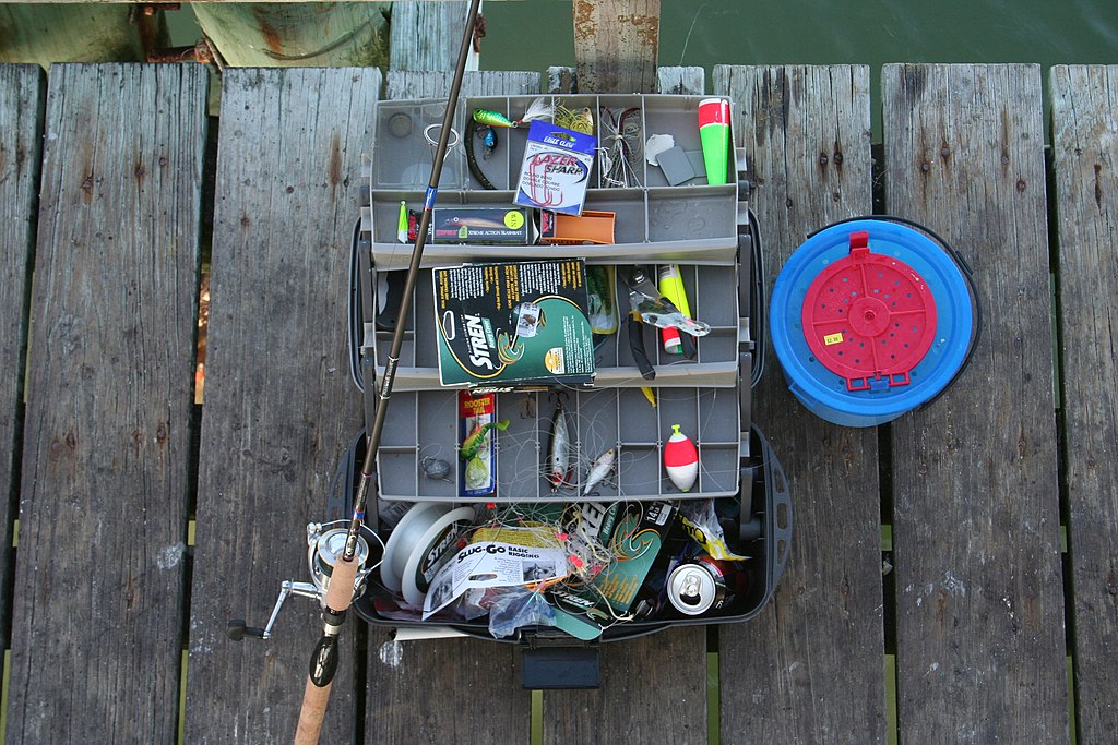 Open tackle box on a wooden dock with various fishing lures, hooks, and a rod. Nearby is a blue bait box, conveying a relaxed angling vibe.