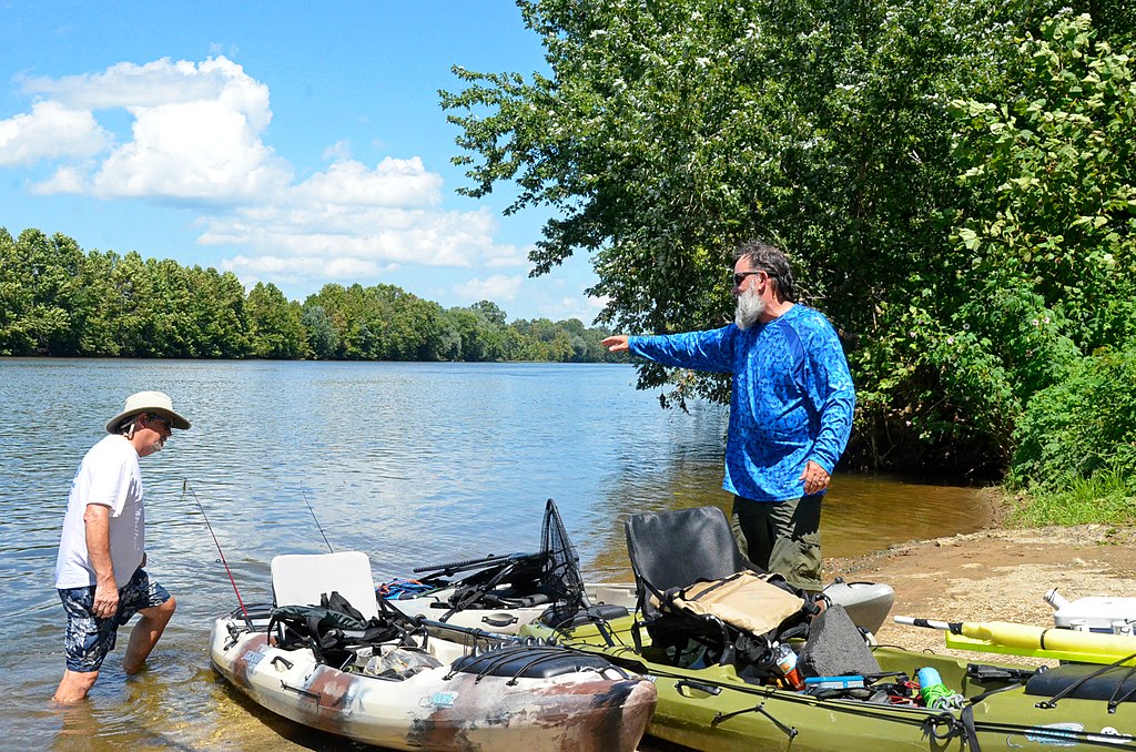 Two men by a riverbank with kayaks, one wading in water and the other pointing. Bright day, lush greenery, and a peaceful, adventurous mood.