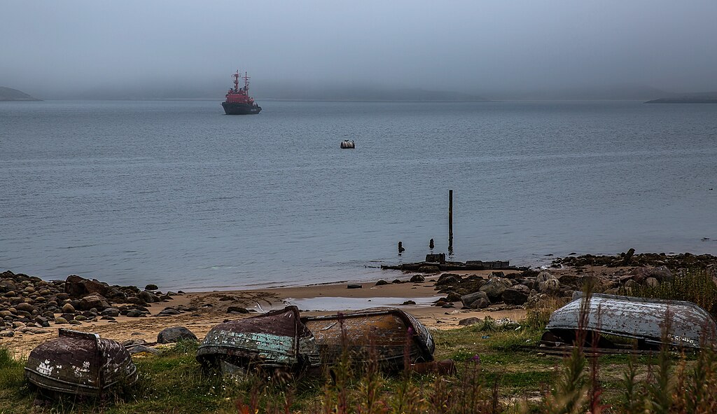 Foggy seascape featuring a red ship in the distance and abandoned boats on a rocky shore in the foreground, conveying a tranquil, melancholic mood.