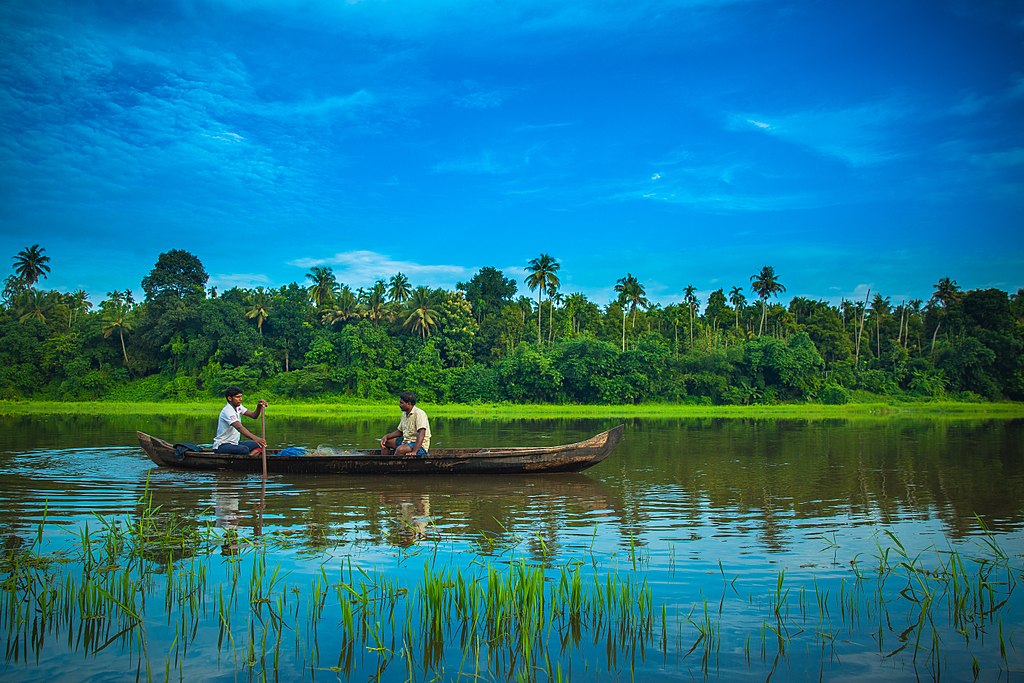 Two people sit in a wooden boat on a calm river, against a backdrop of lush green trees and a vibrant blue sky, conveying a peaceful scene.