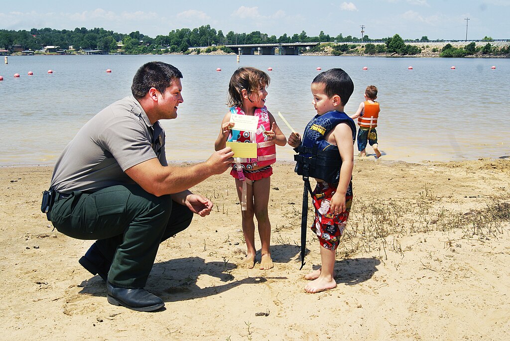 Park ranger kneels on a sandy lakeshore showing a pamphlet to two children in life vests. The background features buoyed water and a distant bridge.