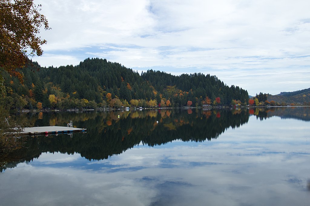 A serene lake reflects autumn trees and a cloudy sky, with a small dock on the left. The vibrant colors and calm water convey tranquility.