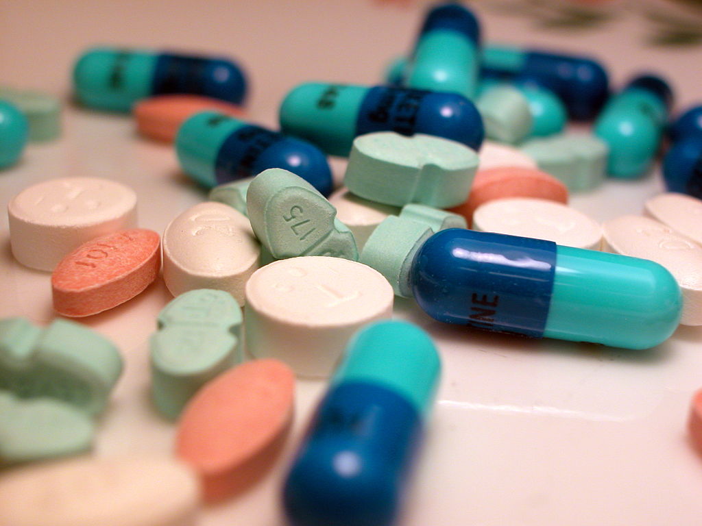 A mix of colorful pills scattered on a surface. Includes blue capsules, white round pills, aqua and peach tablets, evoking a clinical atmosphere.