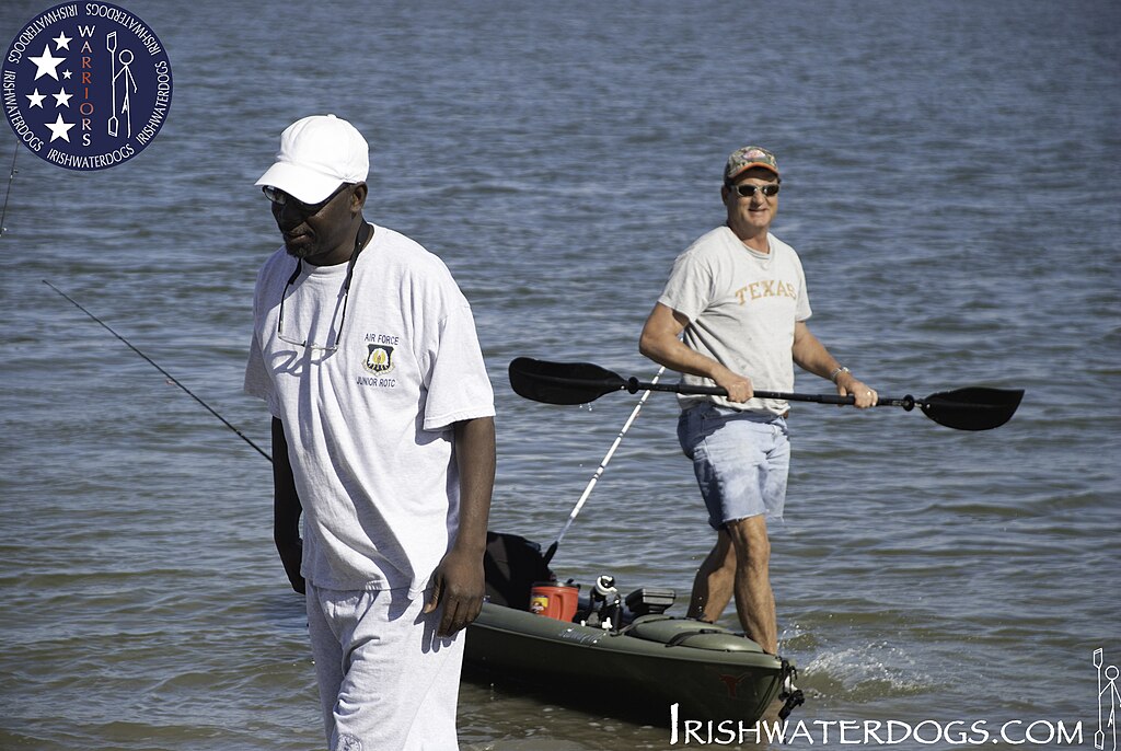 Two men enjoying a day at the water's edge. One man in a white outfit walks with a fishing rod, while the other stands on a kayak, smiling.