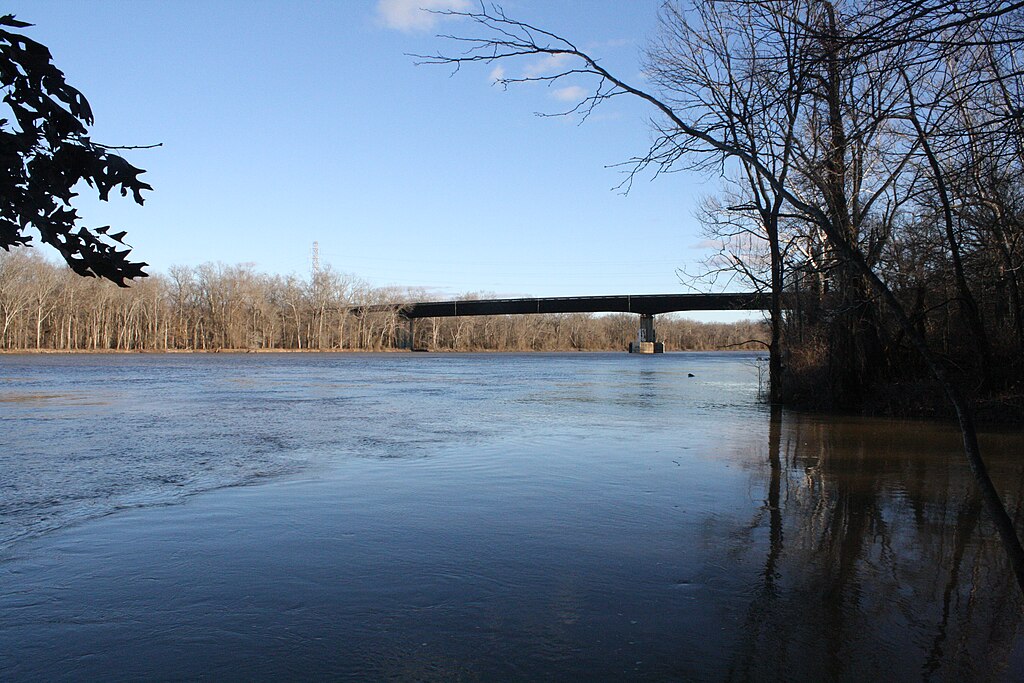 Calm river scene with a bridge spanning across. Bare trees line the banks under a clear blue sky, conveying a peaceful, wintery atmosphere.