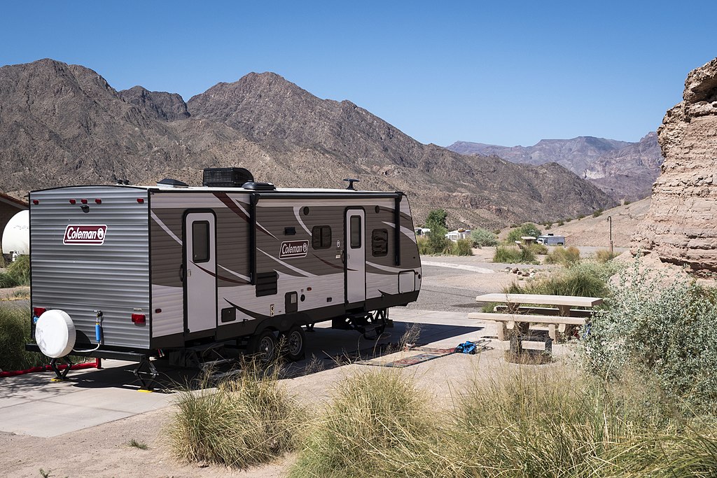 A camper trailer is parked in a desert campsite, surrounded by rocky mountains and sparse vegetation under a clear blue sky, conveying a sense of adventure.