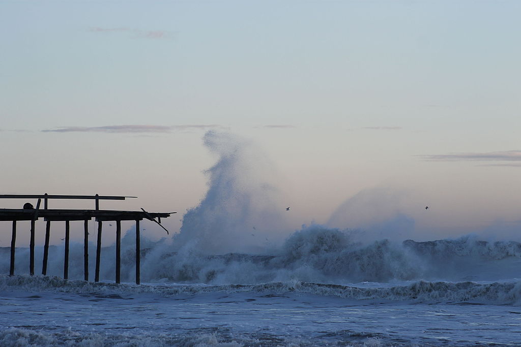 Raging ocean waves crash against a weathered pier at dusk, with silhouettes of birds in flight, creating a dramatic and powerful seascape.