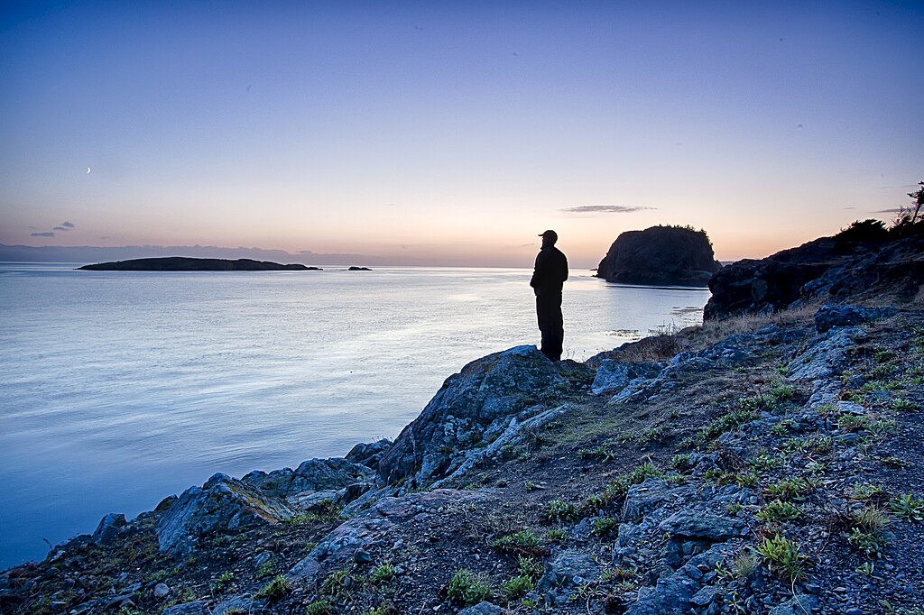 Silhouette of a person standing on a rocky cliff overlooking a calm sea at sunset. The sky is gradient from deep blue to soft pink, evoking tranquility.