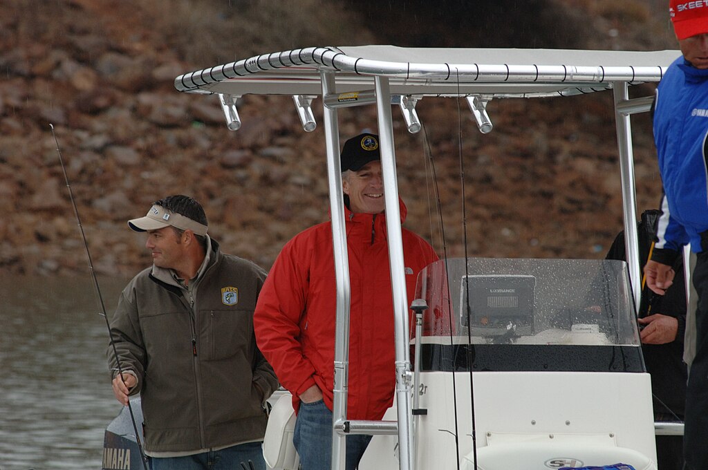 Two men are on a boat during a light rain. One in a red jacket and black cap is smiling, while the other in a gray jacket looks sideways.
