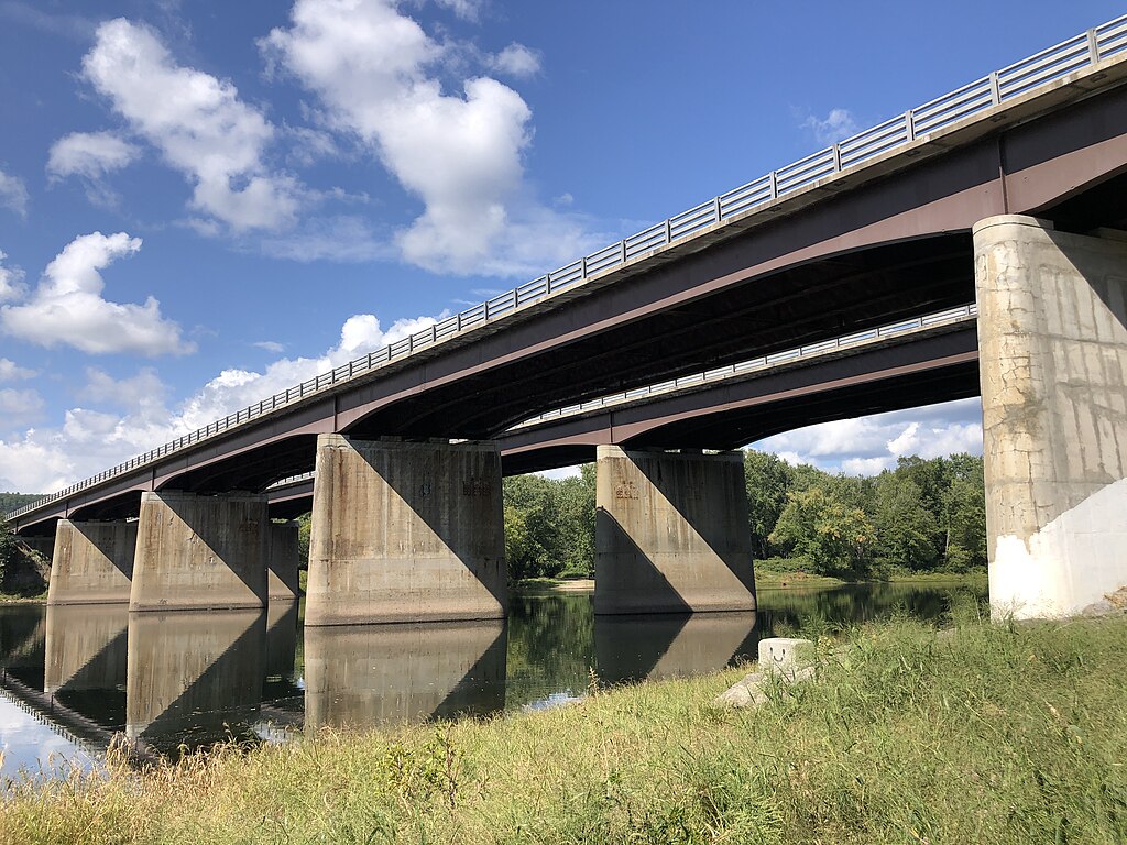 Broad concrete bridge with metal railings spans over a calm river, reflecting its structure. Green banks and trees under a clear, partly cloudy sky.
