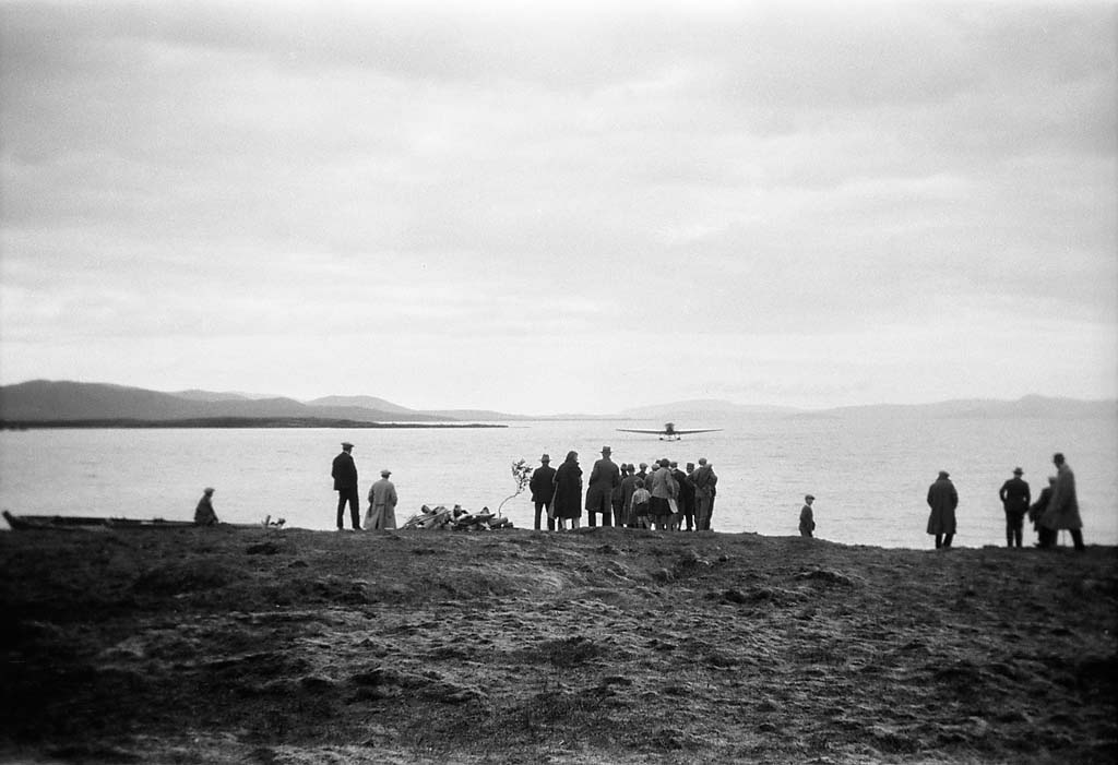 A black-and-white photo shows a group of people on a shoreline watching a small airplane land on a calm lake. The overcast sky and distant hills add a serene mood.