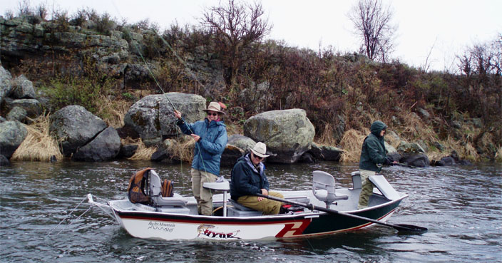 Three people fishing from a small boat on a rocky river with bushes and rocks in the background. One person holds a fishing rod. Overcast day.