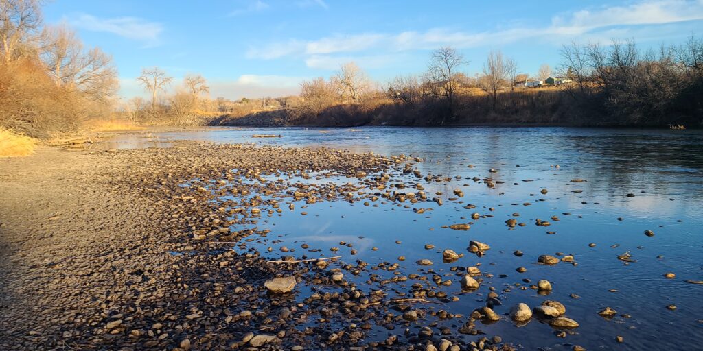 A tranquil river bends gently under a bright blue sky, bordered by bare trees and scattered stones. The serene scene reflects the stillness of winter.