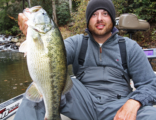 Man in a beanie and outdoor gear holds a large fish on a boat, set against a forested river backdrop. The scene conveys a sense of accomplishment.