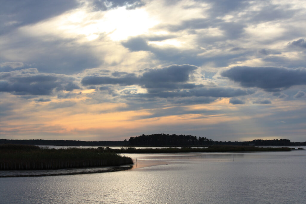 Tranquil sunset scene over a calm waterway with dark silhouetted islands, gently rippling water, and a dramatic sky of scattered clouds and warm hues.