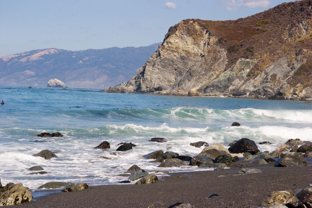Rocky shoreline with waves crashing on the beach, backed by rugged cliffs under a clear blue sky. The scene conveys a sense of serene wilderness.