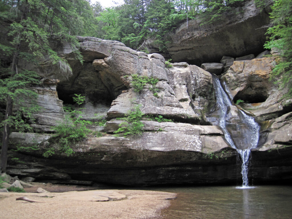 Rocky waterfall amidst lush greenery, cascading into a calm pool below. The scene conveys tranquility and natural beauty, with cave formations above.