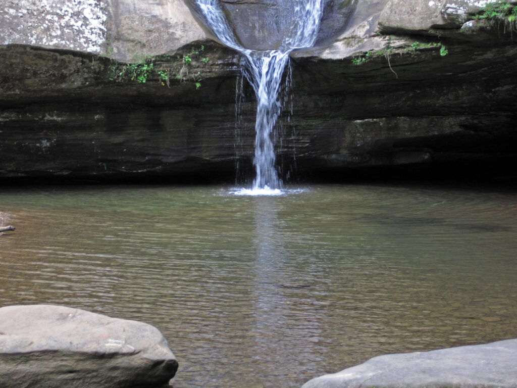 A serene waterfall cascades down a rocky ledge into a tranquil, shallow pool surrounded by moss-covered stones and lush greenery, conveying calm and freshness.