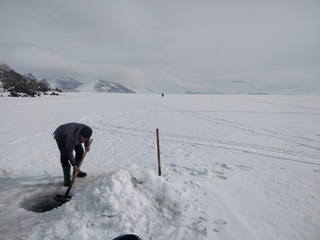 Man stands on a snowy landscape, digging a hole in the ice. Distant mountains and a cloudy sky create a cold, serene scenery.