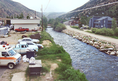 A scenic view of a narrow river flowing through a small town, bordered by lush greenery and rocky banks, with parked cars and buildings nearby under an overcast sky.
